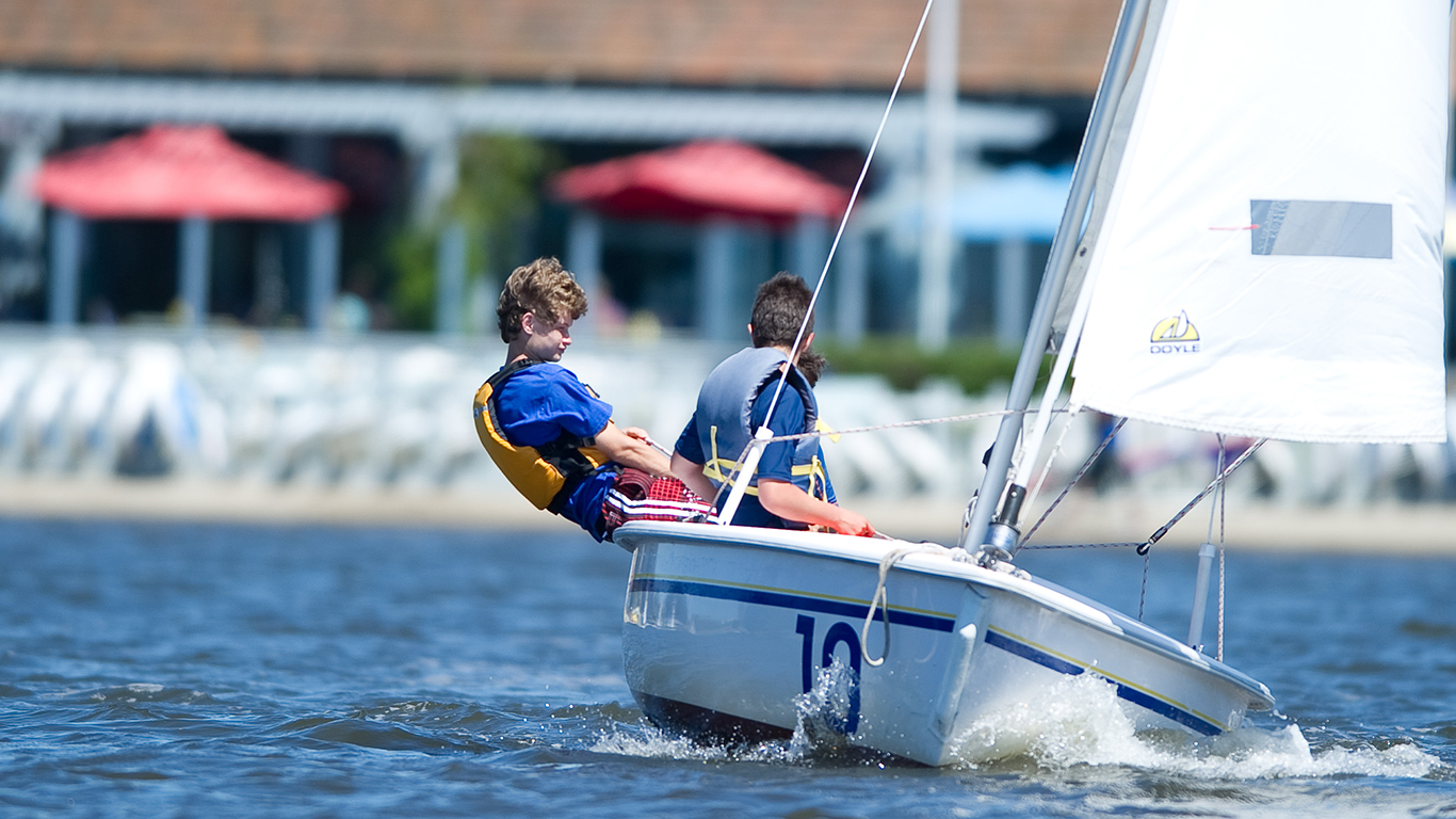 Two boys in a sailboat boat on shoreline lake.